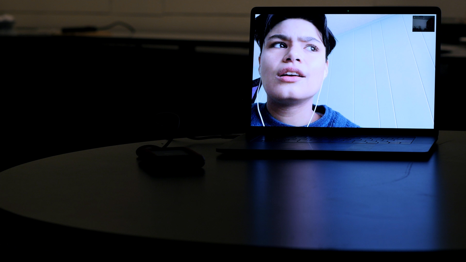 Laptop on a table in a dark room. Laptop screen is bright and features a person on a video call.